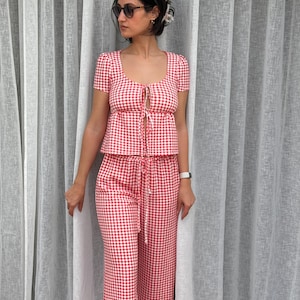 May include: A two-piece set featuring a red and white gingham pattern. The top has short sleeves, a scoop neckline, and a front tie closure. The matching pants feature a high-waisted design with a drawstring waist and a straight-leg silhouette.