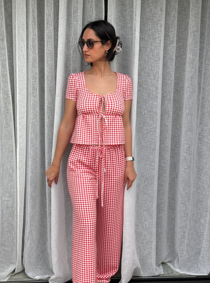 May include: A two-piece set featuring a red and white gingham pattern. The top has short sleeves, a scoop neckline, and a front tie closure. The matching pants feature a high-waisted design with a drawstring waist and a straight-leg silhouette.