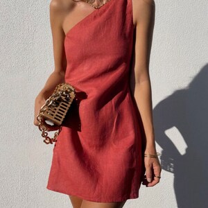 May include: A one-shoulder, terracotta-colored linen dress with a fitted bodice and a slightly flared skirt. The dress is made of a lightweight, breathable fabric and has a relaxed fit.