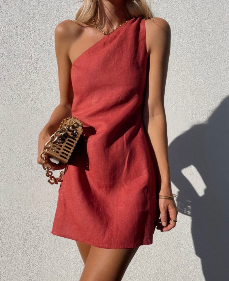 May include: A one-shoulder, terracotta-colored linen dress with a fitted bodice and a slightly flared skirt. The dress is made of a lightweight, breathable fabric and has a relaxed fit.