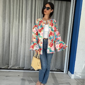 May include: A colorful floral print jacket with bell sleeves. The jacket features a vibrant mix of red, pink, blue, and yellow flowers on a light background. The jacket is paired with blue jeans and a straw handbag.