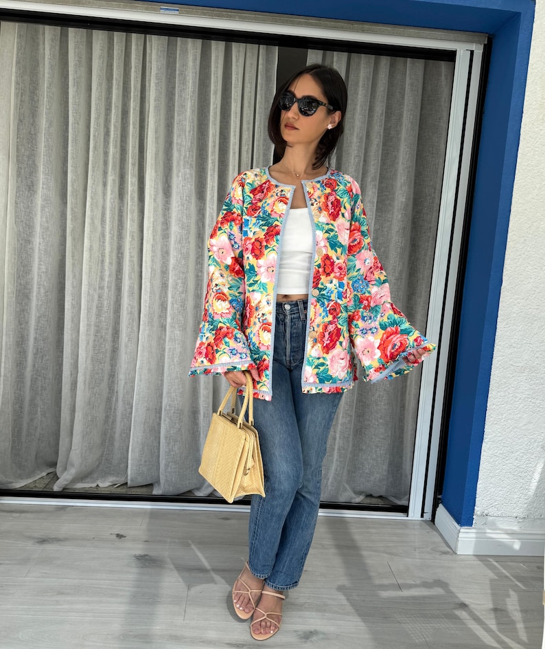May include: A colorful floral print jacket with bell sleeves. The jacket features a vibrant mix of red, pink, blue, and yellow flowers on a light background. The jacket is paired with blue jeans and a straw handbag.