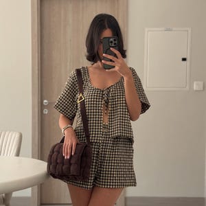 K&ouml;nnte beinhalten: Ein zweiteiliges Set bestehend aus einem kurz&auml;rmeligen Oberteil mit Bindeband vorne und einer passenden Shorts, beide mit einem schwarz-beigen Vichy-Muster. Eine dunkelbraune, rechteckige Umh&auml;ngetasche mit Webstruktur und goldener Schnalle.