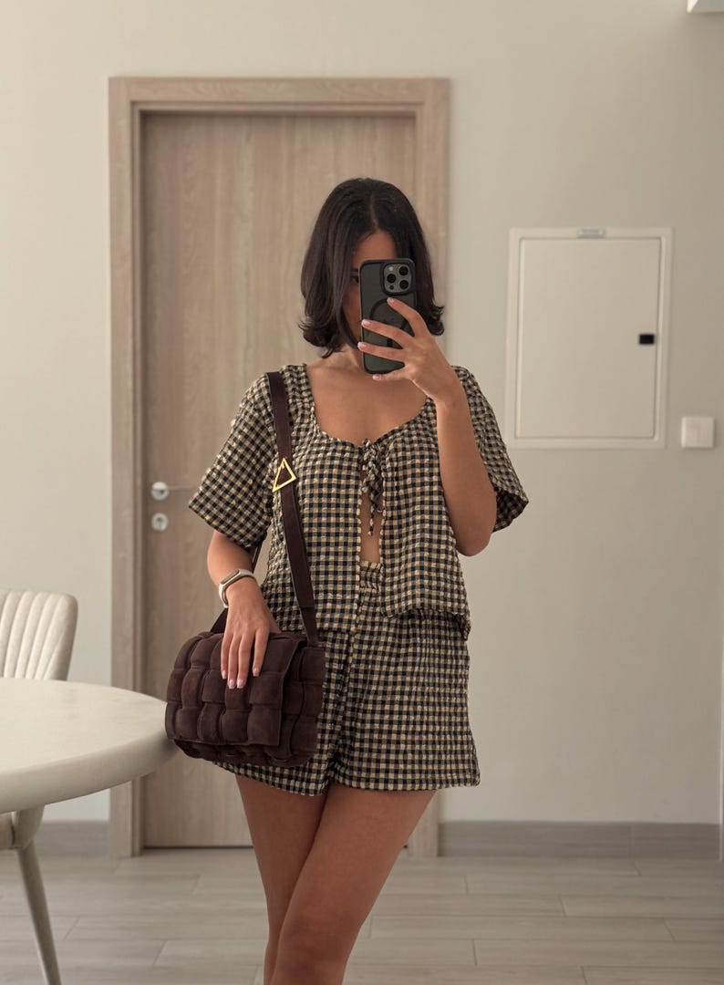 K&ouml;nnte beinhalten: Ein zweiteiliges Set bestehend aus einem kurz&auml;rmeligen Oberteil mit Bindeband vorne und einer passenden Shorts, beide mit einem schwarz-beigen Vichy-Muster. Eine dunkelbraune, rechteckige Umh&auml;ngetasche mit Webstruktur und goldener Schnalle.