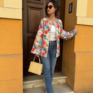 May include: A floral print jacket with a light blue trim and bell sleeves. The jacket features a vibrant pattern of red, pink, and blue flowers. A yellow handbag is held in the hand. The person is wearing blue jeans.