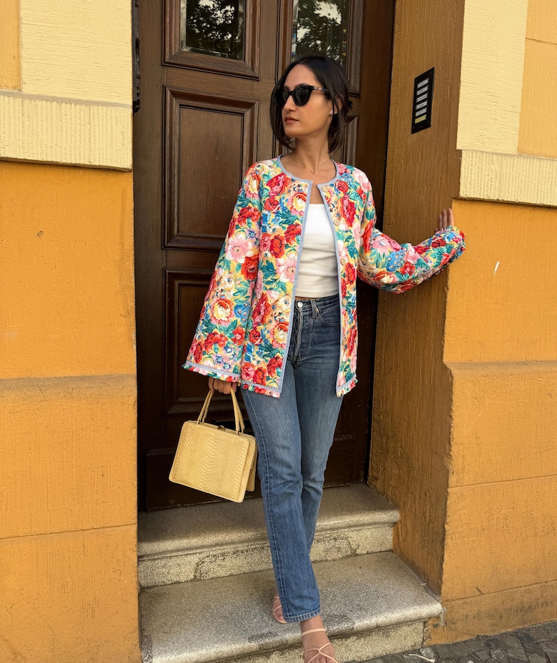 May include: A floral print jacket with a light blue trim and bell sleeves. The jacket features a vibrant pattern of red, pink, and blue flowers. A yellow handbag is held in the hand. The person is wearing blue jeans.