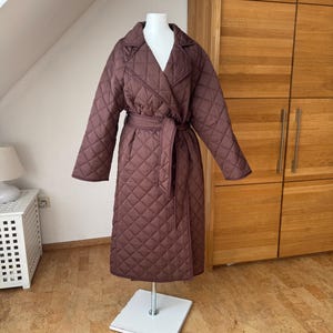 Op de afbeelding: Een lange, gewatteerde bruine jas met een wikkelriem, gepresenteerd op een mannequin. De jas heeft een ruitpatroon, een reverskraag en lange mouwen. De kleur is een diepe bruintint.