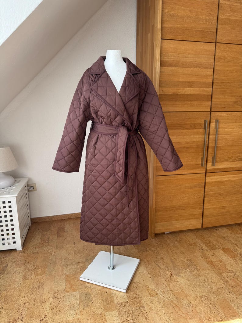 Op de afbeelding: Een lange, gewatteerde bruine jas met een wikkelriem, gepresenteerd op een mannequin. De jas heeft een ruitpatroon, een reverskraag en lange mouwen. De kleur is een diepe bruintint.