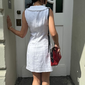 May include: A sleeveless white mini dress featuring a wide collar with a ruffled edge and vertical seam detailing. The dress is paired with a structured handbag that has a red suede body, black leather trim, and a long black shoulder strap.