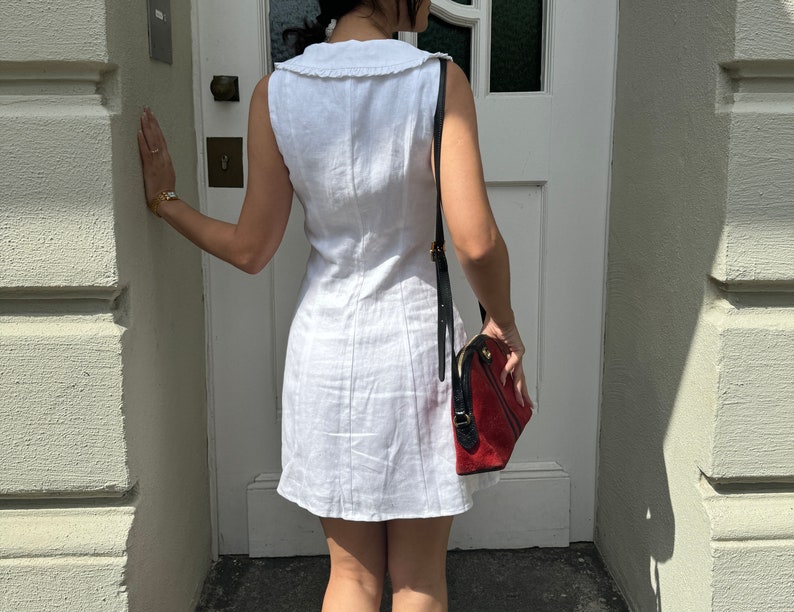 May include: A sleeveless white mini dress featuring a wide collar with a ruffled edge and vertical seam detailing. The dress is paired with a structured handbag that has a red suede body, black leather trim, and a long black shoulder strap.