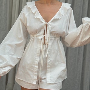 May include: A white two-piece set consisting of a long-sleeved top and matching shorts. The top features a deep V-neckline with ruffled trim, voluminous sleeves with gathered cuffs, and two thin tie closures at the center front over a cinched waist.