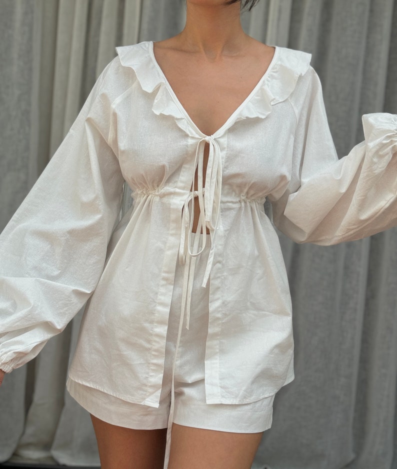 May include: A white two-piece set consisting of a long-sleeved top and matching shorts. The top features a deep V-neckline with ruffled trim, voluminous sleeves with gathered cuffs, and two thin tie closures at the center front over a cinched waist.