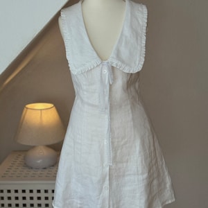 May include: A sleeveless white mini dress featuring a wide collar with ruffled edges, a deep V-neckline, and a front button closure. The dress includes a delicate lace trim detail extending from the collar down the center front placket.
