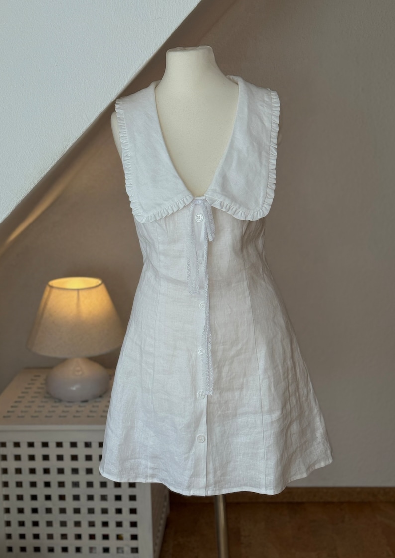 May include: A sleeveless white mini dress featuring a wide collar with ruffled edges, a deep V-neckline, and a front button closure. The dress includes a delicate lace trim detail extending from the collar down the center front placket.