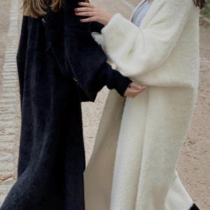 Op de afbeelding: Twee vrouwen dragen lange, pluche cardigans. E&eacute;n cardigan is zwart en de andere is wit.