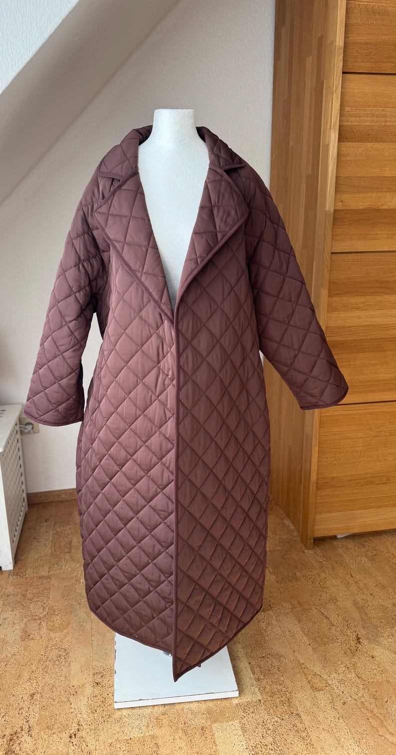 Op de afbeelding: Een lange, gewatteerde jas in een diepbruine tint. De jas heeft een klassieke kraag en een ruitvormig patroon. Het wordt getoond op een mannequin, die de volledige lengte en de getailleerde pasvorm laat zien. De jas lijkt gemaakt van een zacht, warm materiaal.