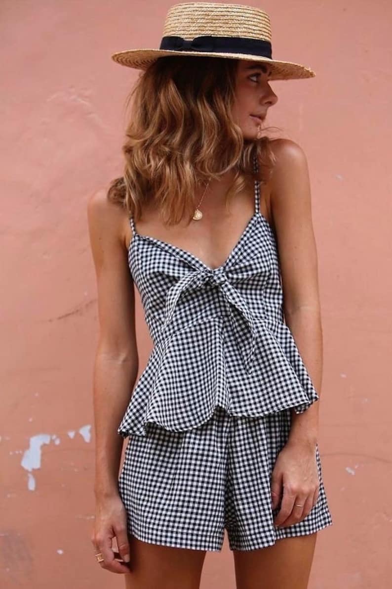 May include: A woman wearing a black and white gingham print top and shorts with a straw hat. The top has a bow at the front.