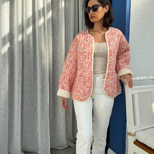 以下が含まれることがあります： ピンクと赤の花柄のキルティングジャケット、クリーム色のトリム付き。 ジャケットはフロントオープンで、ゆったりとしたフィット感です。 白いジーンズと明るい色のトップスを着ています。 ジャケットの長さは約60cmです。