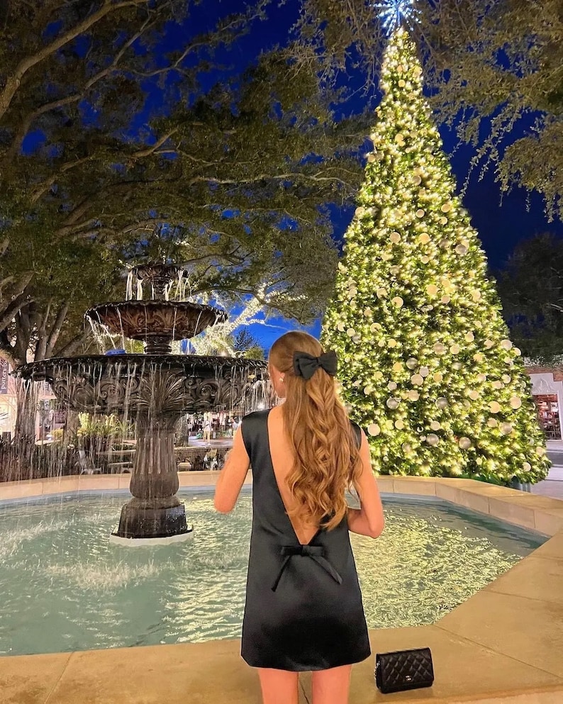 May include: A woman wearing a black sleeveless dress with a low back and a bow at the waist stands in front of a fountain and a decorated Christmas tree. The fountain is made of stone and has a large basin filled with water. The Christmas tree is decorated with white lights and ornaments.