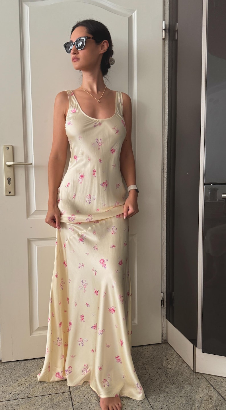 May include: A woman wearing a light yellow maxi dress with a floral pattern. The dress has a scoop neck and thin straps. The woman is wearing sunglasses and a necklace.