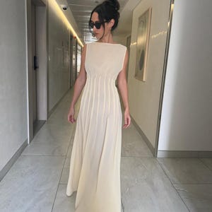 May include: A sleeveless, floor-length cream-colored dress with a pleated skirt and a fitted bodice. The dress has a boat neckline and a flowing silhouette. The person is wearing sunglasses and has their hair up in a bun.