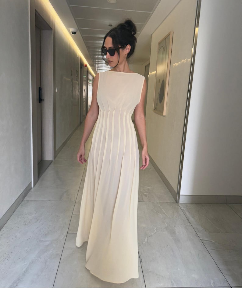 May include: A sleeveless, floor-length cream-colored dress with a pleated skirt and a fitted bodice. The dress has a boat neckline and a flowing silhouette. The person is wearing sunglasses and has their hair up in a bun.