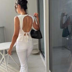 May include: A white sleeveless top with a large back cutout and a tie at the waist. The top is paired with white pants. A black woven handbag is held in the right hand. The person is standing near a glass door.