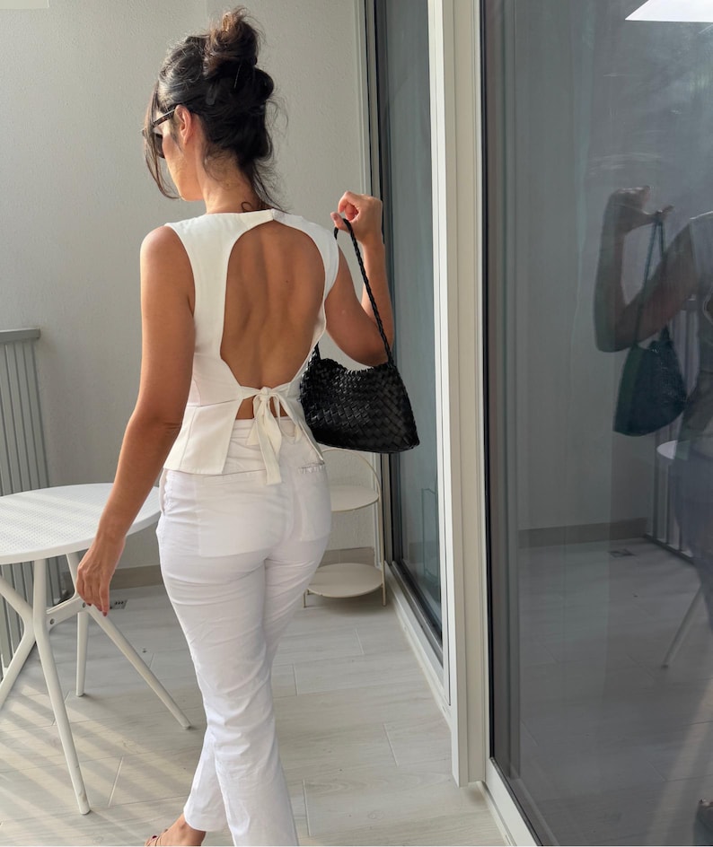 May include: A white sleeveless top with a large back cutout and a tie at the waist. The top is paired with white pants. A black woven handbag is held in the right hand. The person is standing near a glass door.