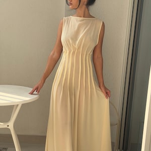 May include: A sleeveless, floor-length, cream-colored dress. The dress features vertical pleats on the bodice and a flowing skirt. The model is wearing sunglasses and standing near a white table and a window.