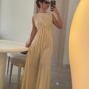May include: A sleeveless, floor-length, cream-colored dress with vertical pleats on the bodice. The dress has a boat neckline and a flowing skirt. The person in the image is wearing sunglasses and holding a phone.