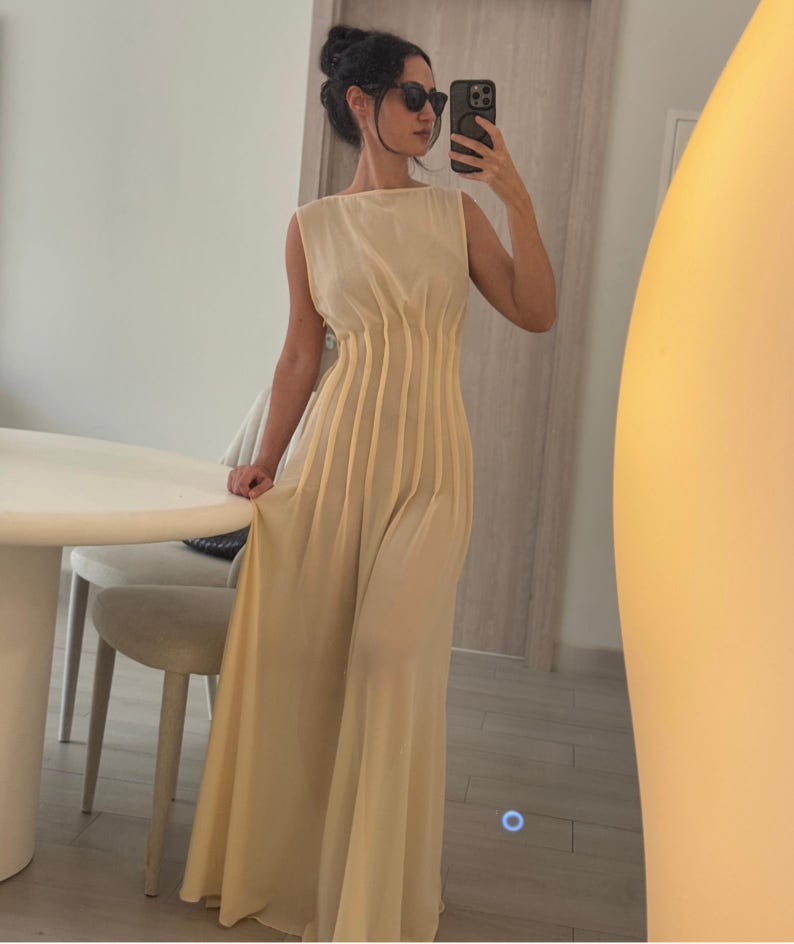May include: A sleeveless, floor-length, cream-colored dress with vertical pleats on the bodice. The dress has a boat neckline and a flowing skirt. The person in the image is wearing sunglasses and holding a phone.
