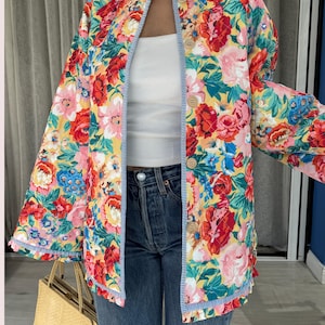 May include: A floral print jacket with a yellow background and red, pink, and blue flowers. The jacket has a blue and white striped trim and wooden buttons. The jacket is open, revealing a white top and blue jeans.