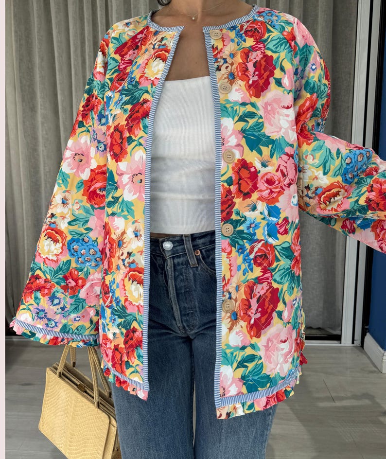 May include: A floral print jacket with a yellow background and red, pink, and blue flowers. The jacket has a blue and white striped trim and wooden buttons. The jacket is open, revealing a white top and blue jeans.