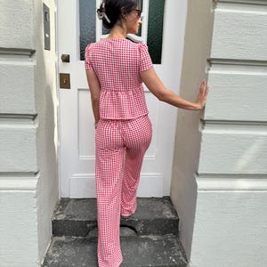 May include: A two-piece set consisting of a short-sleeved peplum top and matching wide-leg trousers, both featuring a red and white gingham check pattern. The top has a fitted bodice with a flared waist, and the trousers have a straight-leg cut.