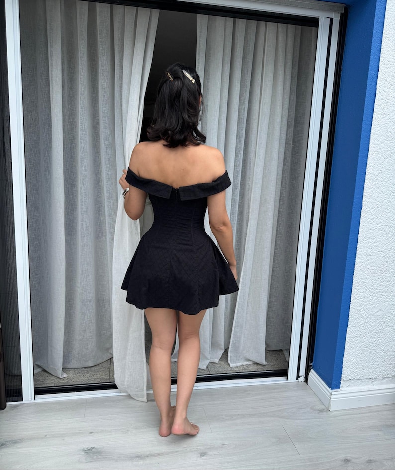 May include: A black off-the-shoulder dress with a fitted bodice and flared skirt. The dress is made of a textured fabric and is worn by a person standing near a window with sheer white curtains. The background includes a blue wall and a white floor.
