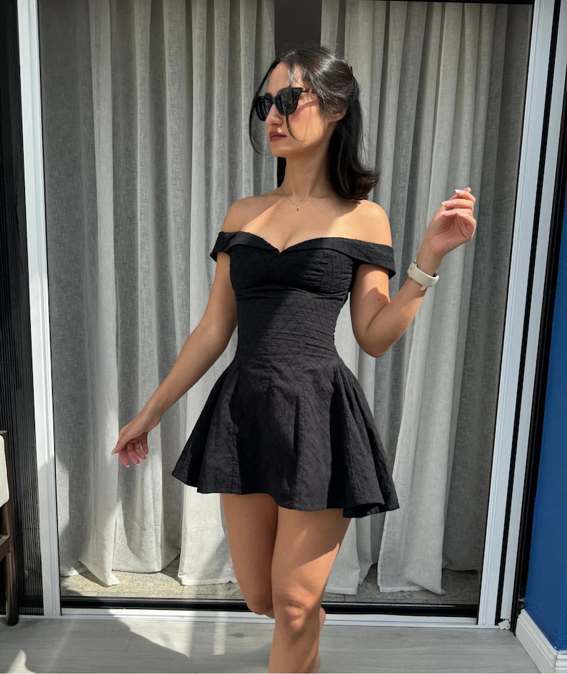 May include: A black off-the-shoulder dress with a fitted bodice and a flared skirt. The dress has a textured pattern and is worn with sunglasses. The dress is a mini dress.