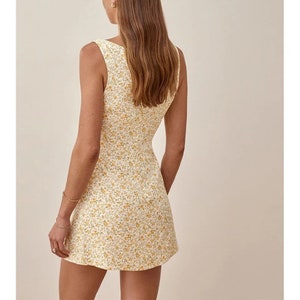 May include: A sleeveless mini dress with a white background and a yellow floral pattern. The dress has a scoop neck and a fitted bodice.