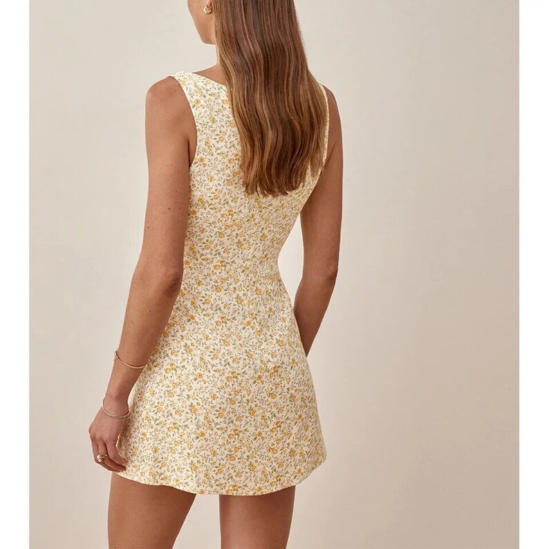 May include: A sleeveless mini dress with a white background and a yellow floral pattern. The dress has a scoop neck and a fitted bodice.