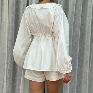 May include: A white long-sleeved blouse featuring a ruffled collar and a cinched waist with a gathered detail, paired with matching white shorts. The blouse has voluminous sleeves with cuffed wrists and a loose, flowing hemline that falls over the shorts.