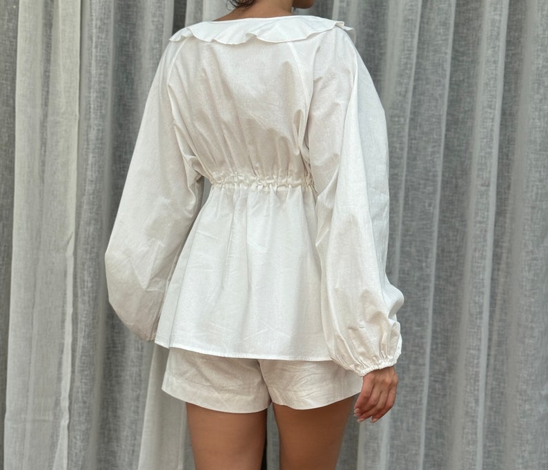 May include: A white long-sleeved blouse featuring a ruffled collar and a cinched waist with a gathered detail, paired with matching white shorts. The blouse has voluminous sleeves with cuffed wrists and a loose, flowing hemline that falls over the shorts.