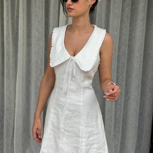 May include: A sleeveless white mini dress featuring a wide, ruffled collar and a front button-down closure. The dress has a fitted bodice and a slightly flared skirt, crafted from a textured fabric with a clean, solid white finish throughout.