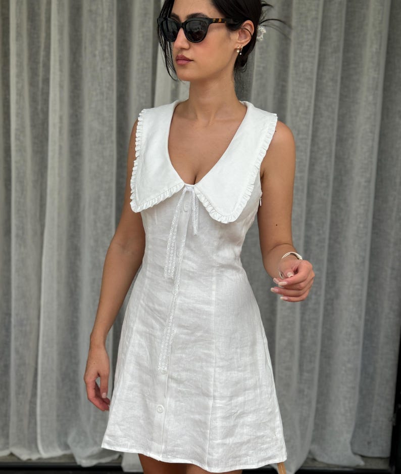 May include: A sleeveless white mini dress featuring a wide, ruffled collar and a front button-down closure. The dress has a fitted bodice and a slightly flared skirt, crafted from a textured fabric with a clean, solid white finish throughout.
