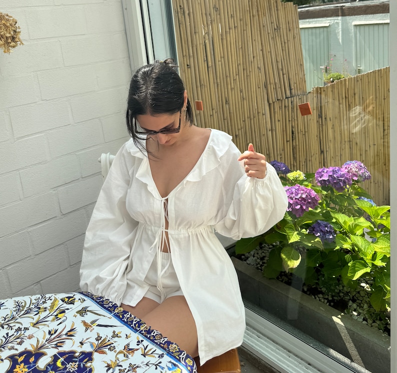 May include: A white long-sleeved blouse featuring a ruffled collar, a deep V-neckline with thin tie closures at the center, and voluminous sleeves. The garment has a cinched waistline and a relaxed, flowing fit that extends to the upper thigh area.