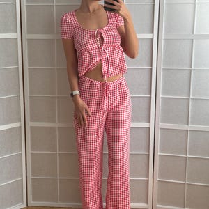 May include: A two-piece set featuring a red and white gingham pattern. The top has short sleeves, a scoop neckline, and a front tie closure. The matching pants feature a high-waisted design with a drawstring waist and a wide-leg silhouette.