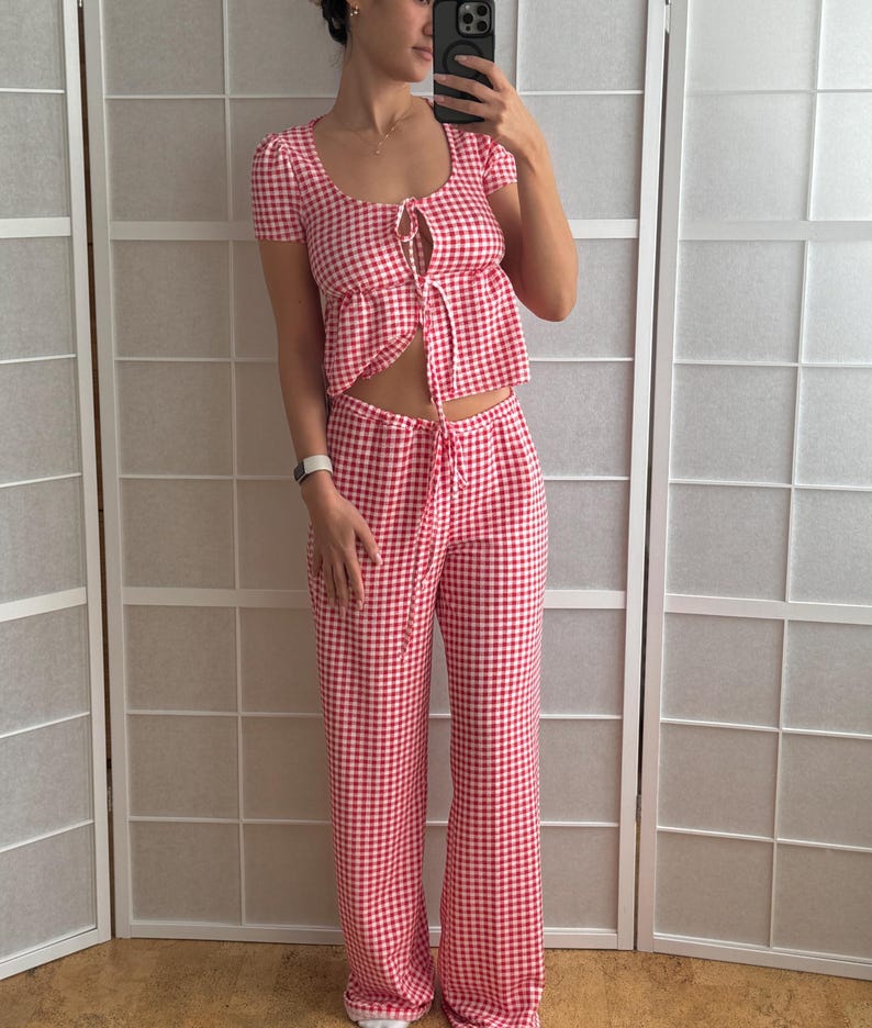 May include: A two-piece set featuring a red and white gingham pattern. The top has short sleeves, a scoop neckline, and a front tie closure. The matching pants feature a high-waisted design with a drawstring waist and a wide-leg silhouette.