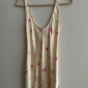 May include: A sleeveless, cream-colored slip dress with a floral print featuring pink and purple flowers. The dress has a scoop neck and a relaxed fit.
