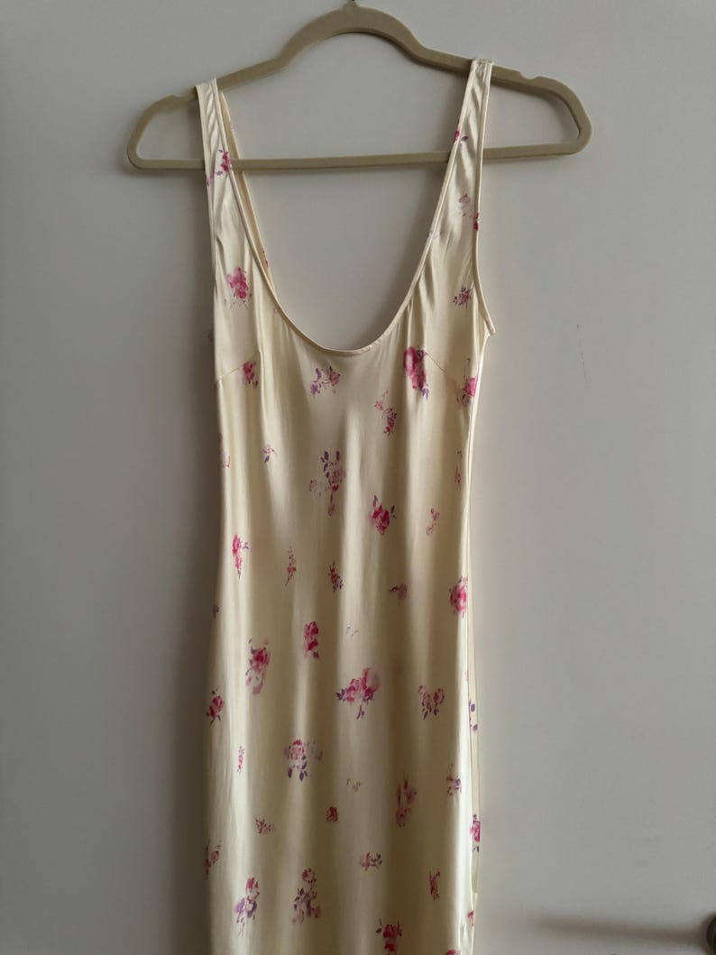 May include: A sleeveless, cream-colored slip dress with a floral print featuring pink and purple flowers. The dress has a scoop neck and a relaxed fit.