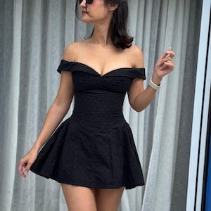 May include: A black off-the-shoulder dress with a fitted bodice and flared skirt. The dress has a textured pattern and is worn with sunglasses.