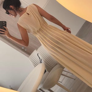 May include: A long, flowing, pale yellow dress with a pleated skirt and a sleeveless design. The dress has a keyhole detail at the back and a cinched waist. The fabric appears lightweight and sheer, creating an elegant silhouette.