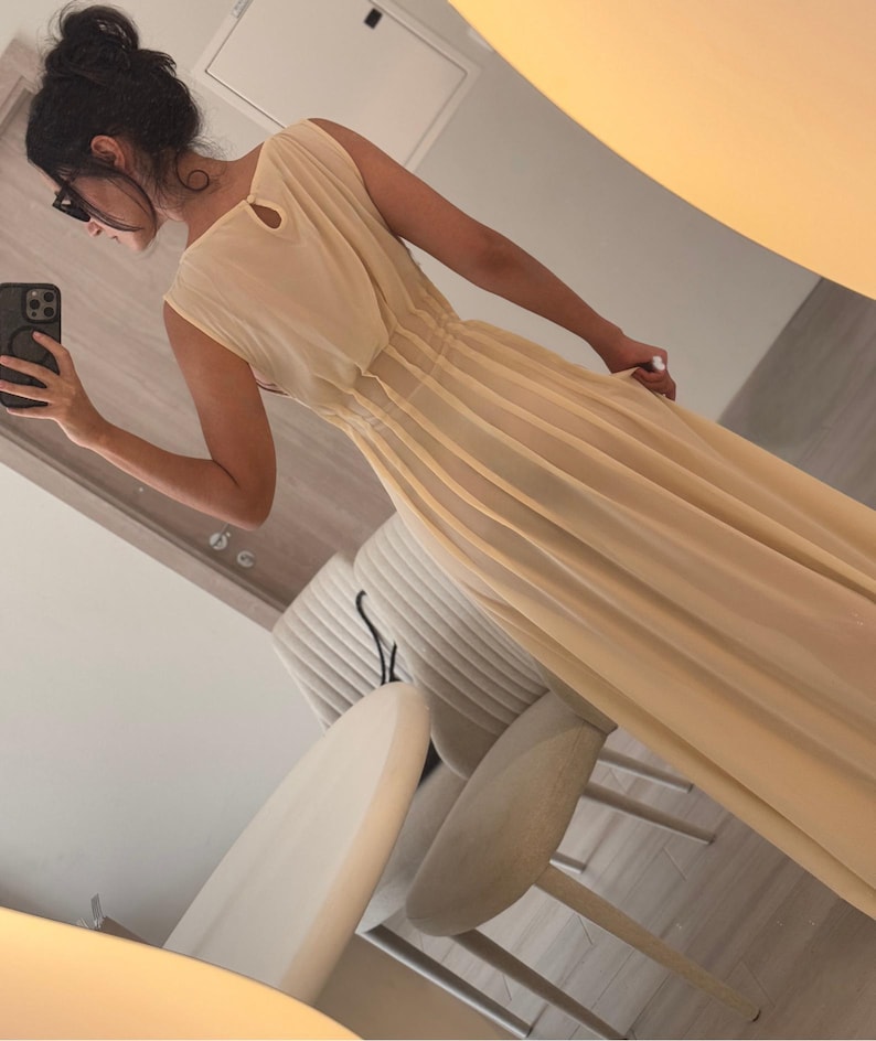 May include: A long, flowing, pale yellow dress with a pleated skirt and a sleeveless design. The dress has a keyhole detail at the back and a cinched waist. The fabric appears lightweight and sheer, creating an elegant silhouette.